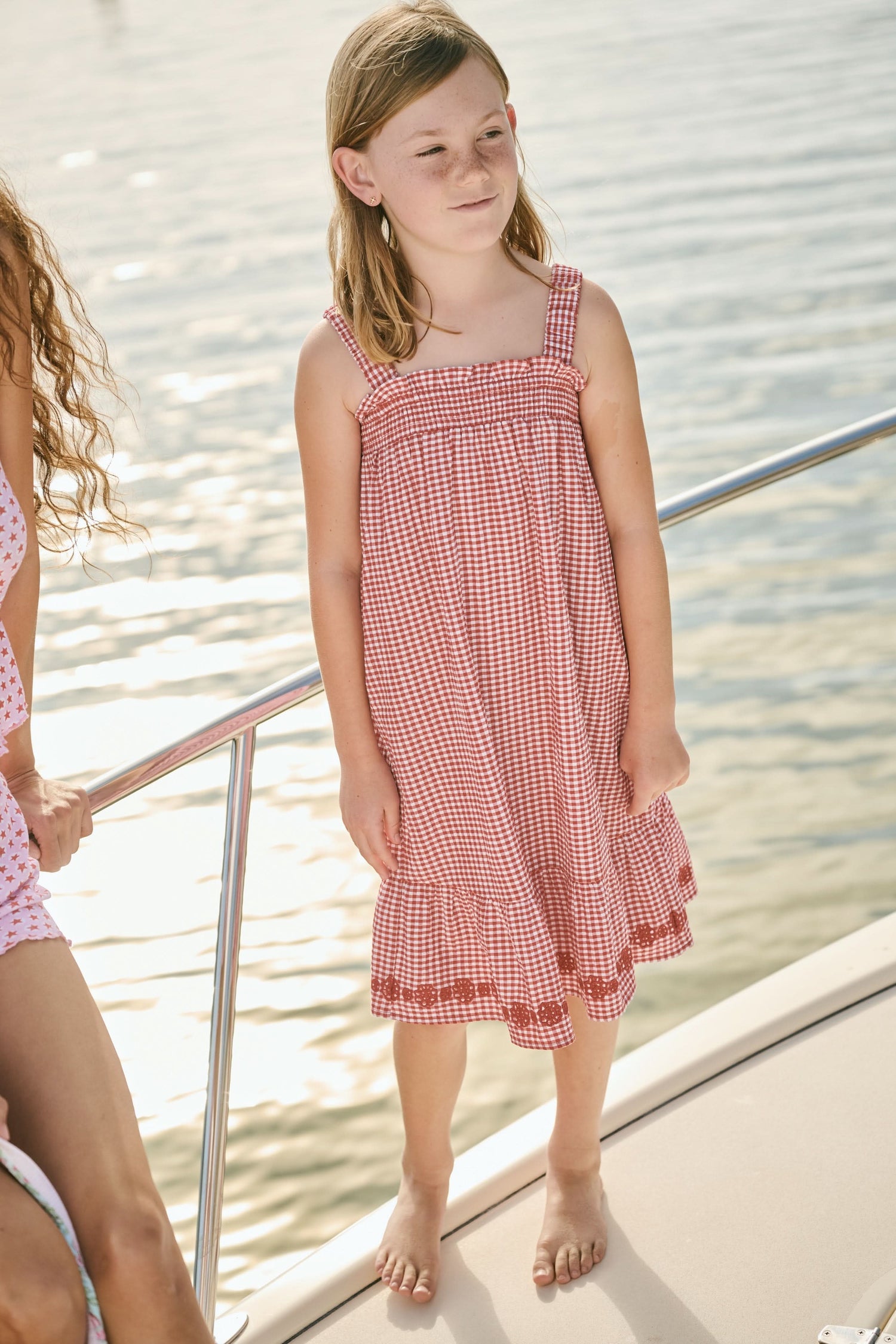 A young girl in the Roller Rabbit Kids Gingham Teagan Dress with an embroidered hem stands barefoot on a boat deck by the water, sunlight shimmering on the surface.