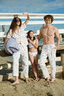 A person stands barefoot in Roller Rabbit’s Men’s Catch of the Day Jon Pant—loose white seersucker pants with a lobsters, fish, and seashells print—and a white t-shirt, one hand tucked into the pants’ pocket.