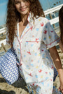 A woman with long curly hair smiles in the Catch of the Day Liza Set by Roller Rabbit, featuring red and blue sea-themed prints. She holds a blue patterned pillow on a sandy beach with a white fence in the background.