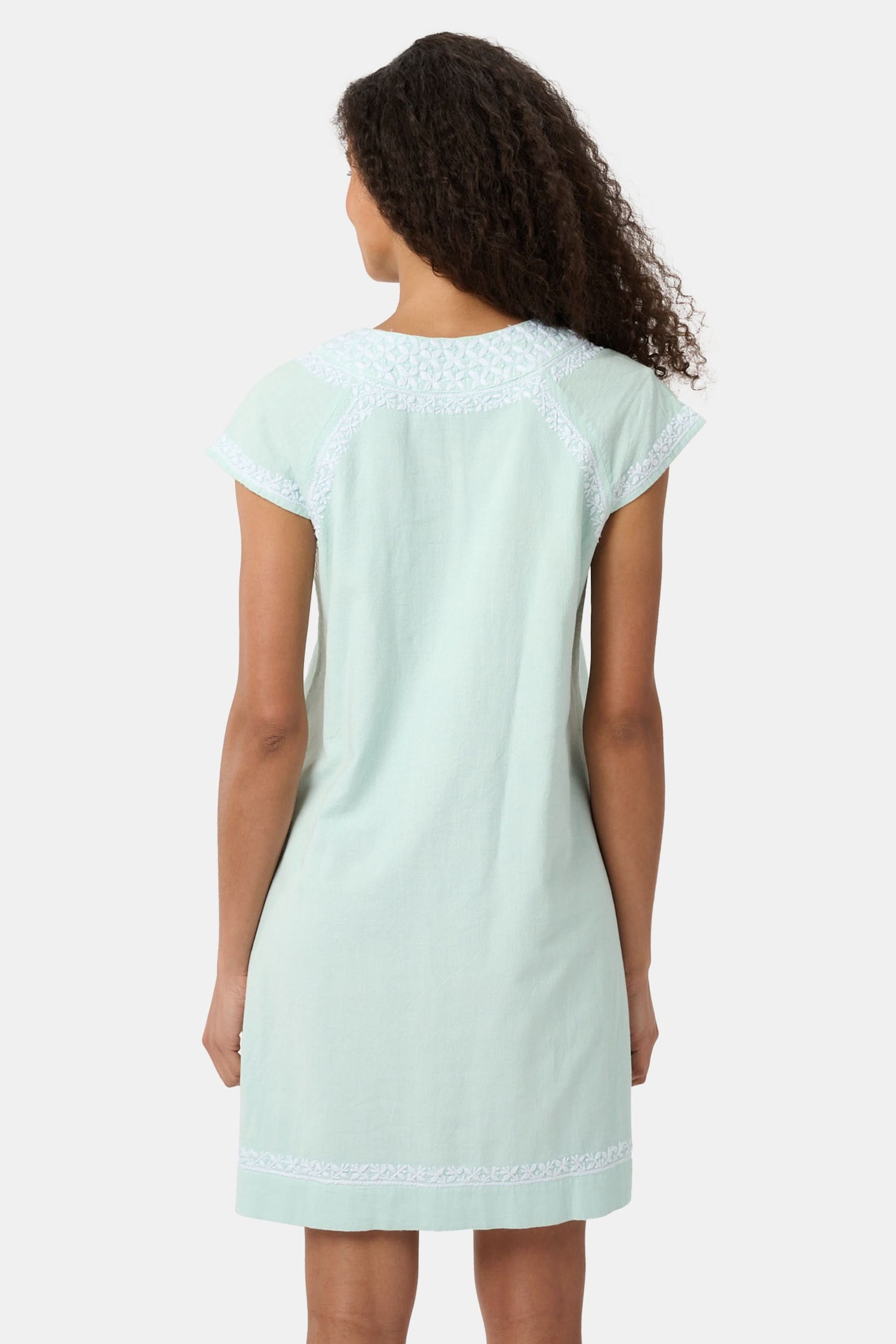 A woman with long curly hair wears the Roller Rabbit Faith Dress, a seaglass-green short-sleeved dress with hand embroidery and a notched neckline, standing against a plain light background.