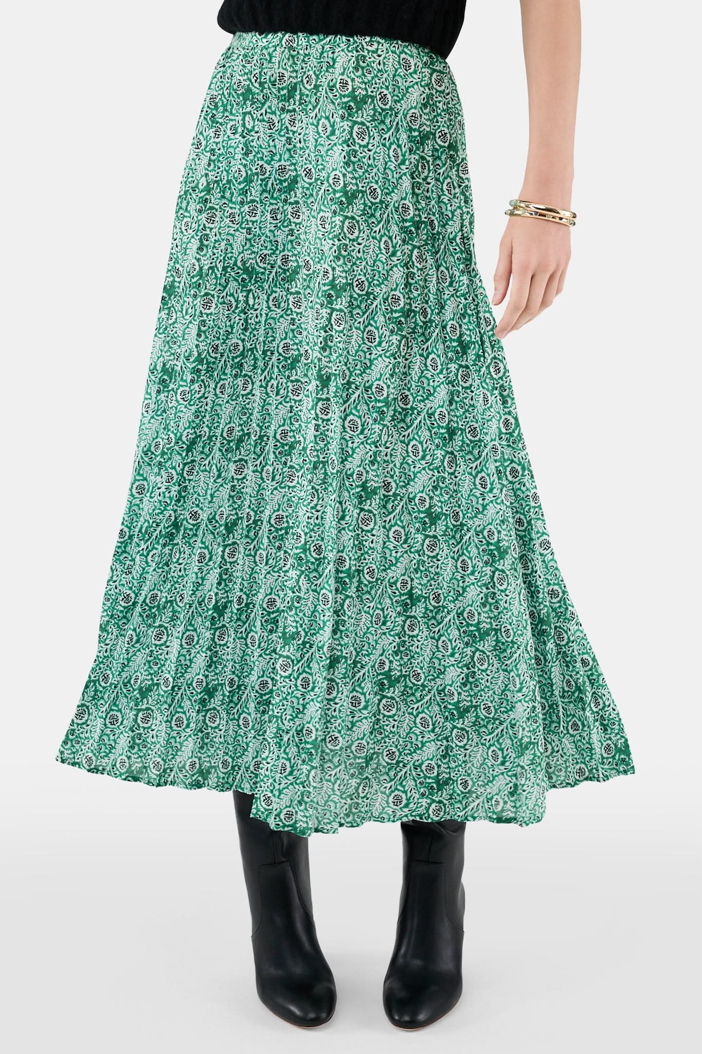 Someone wears the Roller Rabbit Fern Floral Megan Skirt—a green and white pleated maxi—paired with a black top, gold bracelets, and black heeled boots, shown from the waist down against a plain background.