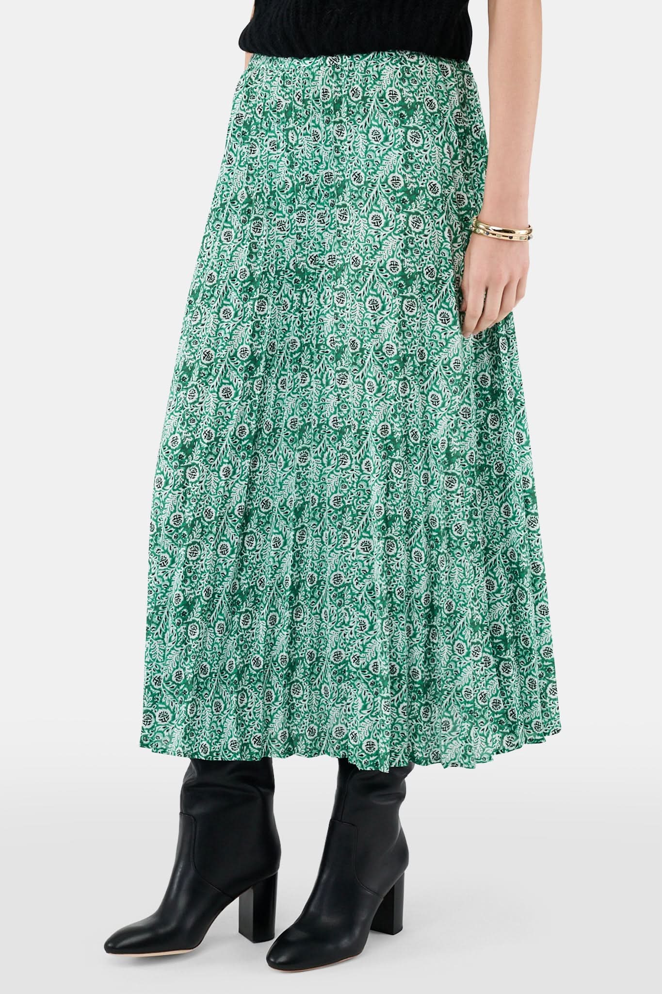 Someone wears the Roller Rabbit Fern Floral Megan Skirt—a green and white pleated maxi—paired with a black top, gold bracelets, and black heeled boots, shown from the waist down against a plain background.