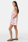 A woman in the Roller Rabbit Fruits D'Ete Keaton Dress with a matching headscarf stands barefoot by a vintage white Volkswagen van, shielding her eyes from the sun. The sunny coastal backdrop highlights the dress's playful print.