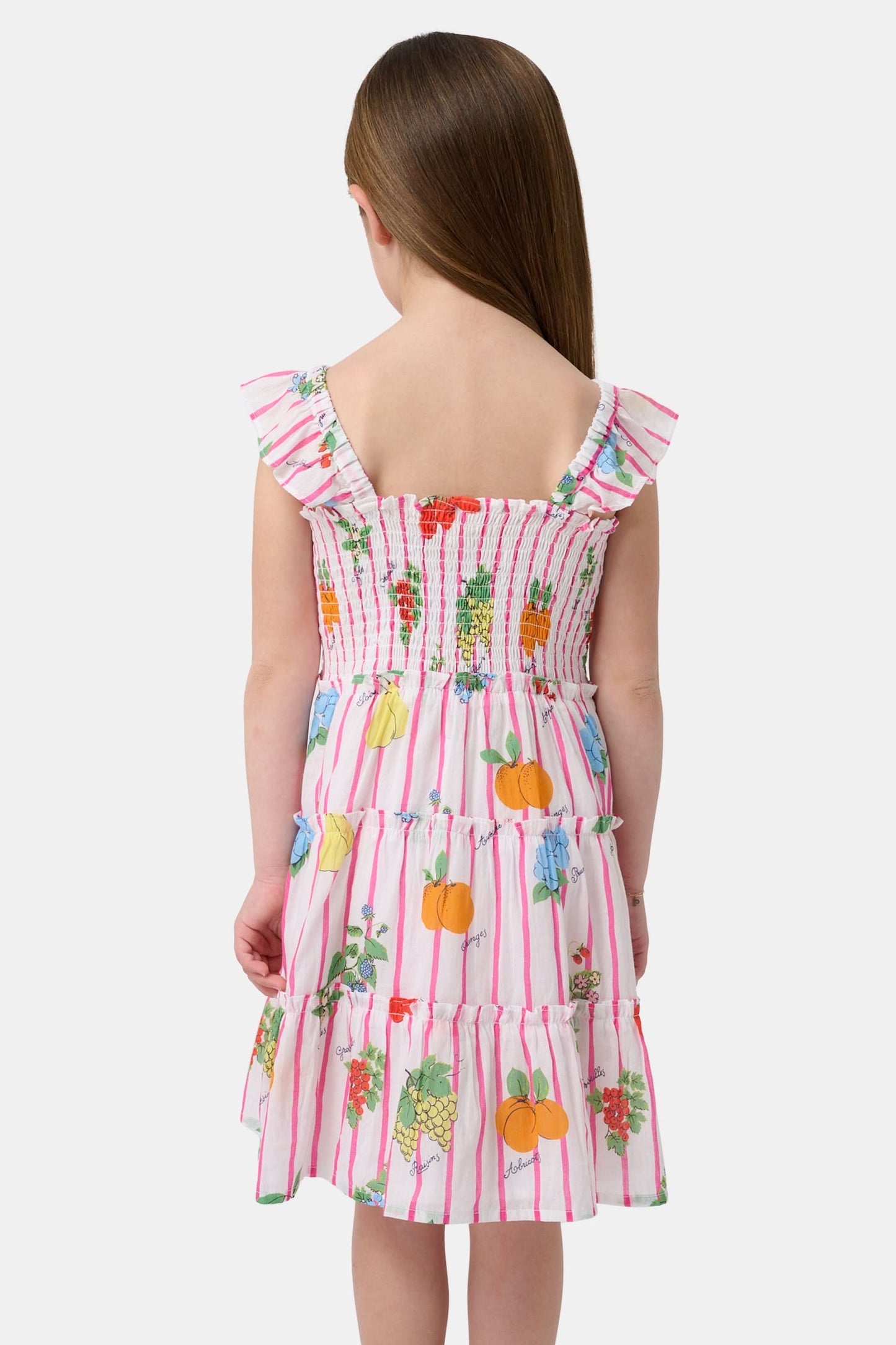 A young girl with straight brown hair wears the Roller Rabbit Kids Fruits D'Ete Ceri Dress, a pink and white smocked sundress with ruffled sleeves, and white sandals, standing and smiling against a light gray background.