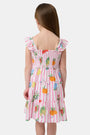A young girl with straight brown hair wears the Roller Rabbit Kids Fruits D'Ete Ceri Dress, a pink and white smocked sundress with ruffled sleeves, and white sandals, standing and smiling against a light gray background.