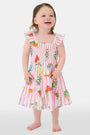 A young girl with straight brown hair wears the Roller Rabbit Kids Fruits D'Ete Ceri Dress, a pink and white smocked sundress with ruffled sleeves, and white sandals, standing and smiling against a light gray background.