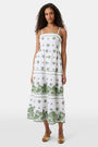 A woman with curly hair wears the Roller Rabbit Fruit Eyelet Selena Dress, a green linen maxi with floral embroidery and thin straps. She stands facing forward in yellow open-toe sandals against a plain white background.
