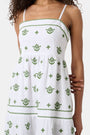 A woman with curly hair wears the Roller Rabbit Fruit Eyelet Selena Dress, a green linen maxi with floral embroidery and thin straps. She stands facing forward in yellow open-toe sandals against a plain white background.