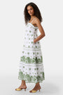 A woman with curly hair wears the Roller Rabbit Fruit Eyelet Selena Dress, a green linen maxi with floral embroidery and thin straps. She stands facing forward in yellow open-toe sandals against a plain white background.