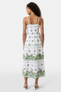 A woman with curly hair wears the Roller Rabbit Fruit Eyelet Selena Dress, a green linen maxi with floral embroidery and thin straps. She stands facing forward in yellow open-toe sandals against a plain white background.