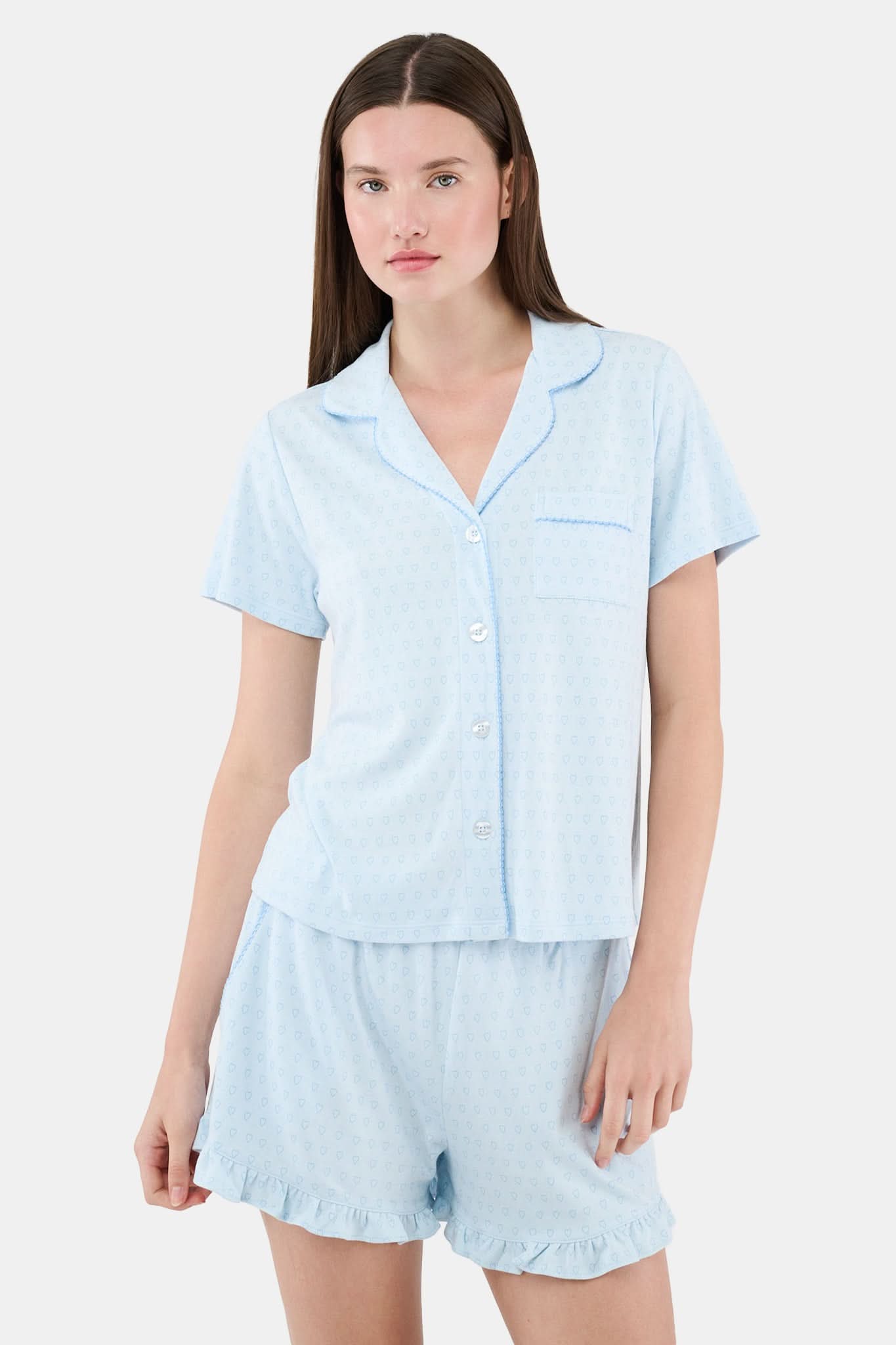 A woman wears Roller Rabbit's Hearts Tonal Wyatt Polo Pajamas—light blue, short-sleeved Pima cotton with a collar and chest pocket—paired with matching ruffled shorts, standing against a plain light background.