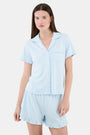 A woman wears Roller Rabbit's Hearts Tonal Wyatt Polo Pajamas—light blue, short-sleeved Pima cotton with a collar and chest pocket—paired with matching ruffled shorts, standing against a plain light background.
