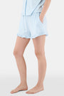 A woman wears Roller Rabbit's Hearts Tonal Wyatt Polo Pajamas—light blue, short-sleeved Pima cotton with a collar and chest pocket—paired with matching ruffled shorts, standing against a plain light background.