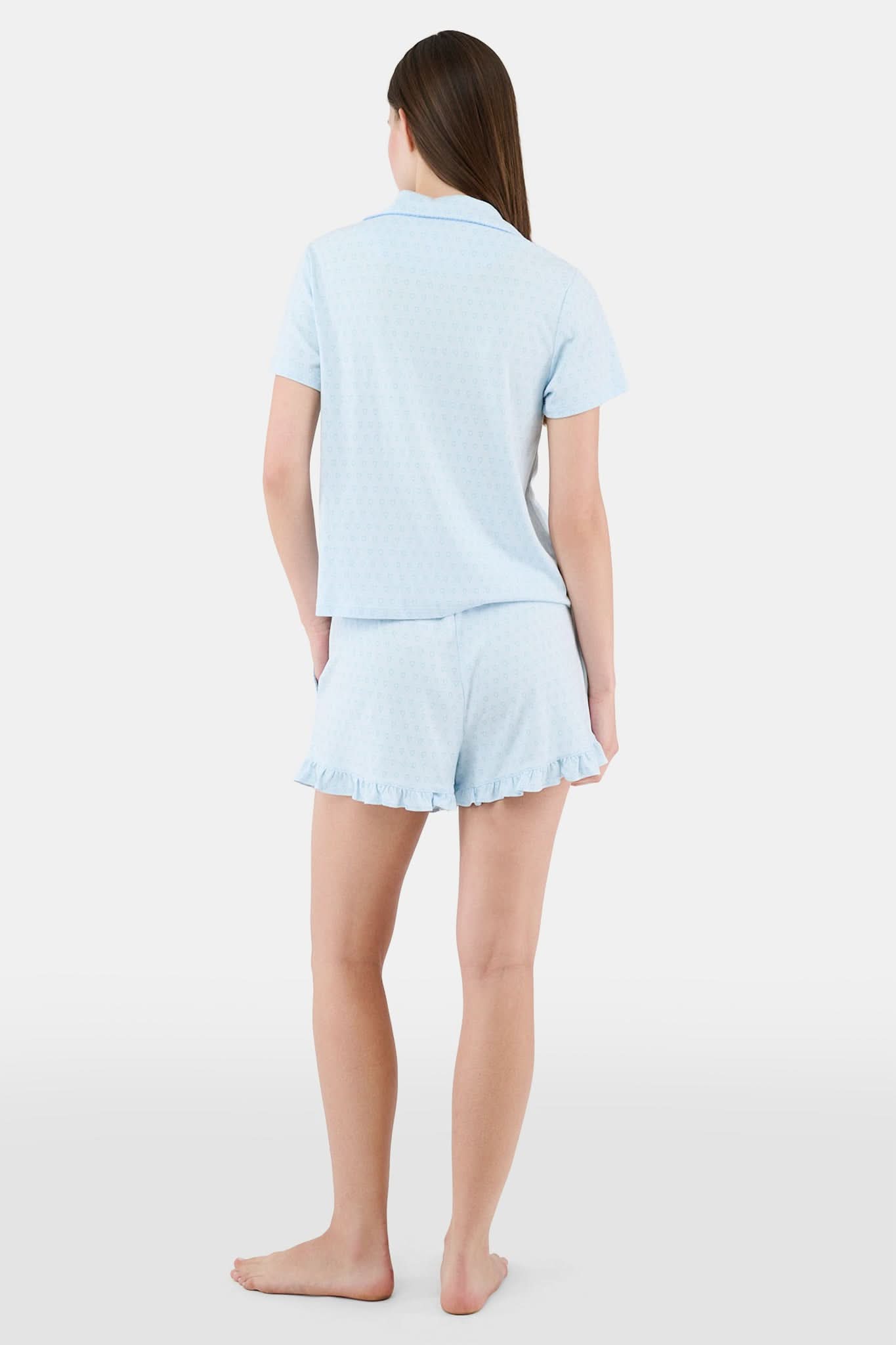 A woman wears Roller Rabbit's Hearts Tonal Wyatt Polo Pajamas—light blue, short-sleeved Pima cotton with a collar and chest pocket—paired with matching ruffled shorts, standing against a plain light background.