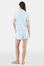 A woman wears Roller Rabbit's Hearts Tonal Wyatt Polo Pajamas—light blue, short-sleeved Pima cotton with a collar and chest pocket—paired with matching ruffled shorts, standing against a plain light background.