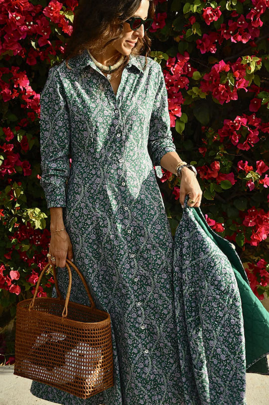 A woman in the Roller Rabbit Suzie Floral Ribbon Darla Dress stands before vibrant pink bougainvillea, holding a woven brown handbag and matching green jacket, accessorized with sunglasses and jewelry.