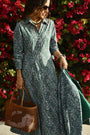 A woman in the Roller Rabbit Suzie Floral Ribbon Darla Dress stands before vibrant pink bougainvillea, holding a woven brown handbag and matching green jacket, accessorized with sunglasses and jewelry.