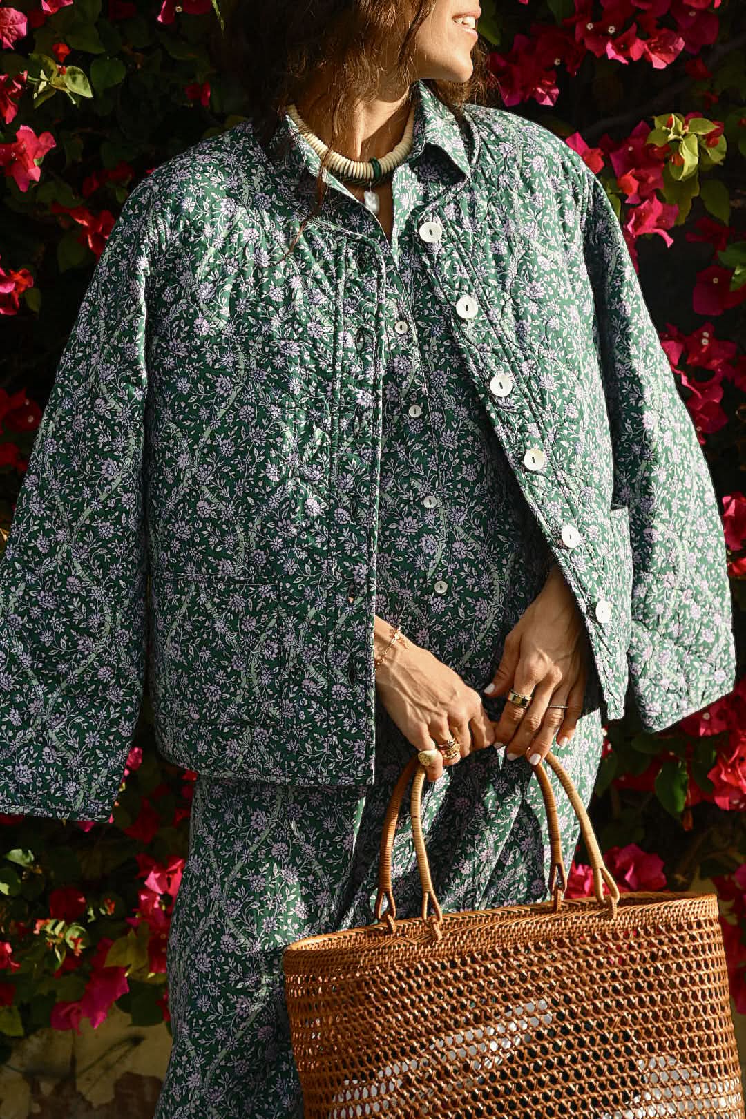 A person wearing the Roller Rabbit Suzie Floral Ribbon Sadie Jacket and a matching skirt stands by blooming pink flowers, holding a brown woven tote. Their face is partially out of frame.