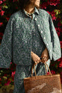 A person wearing the Roller Rabbit Suzie Floral Ribbon Sadie Jacket and a matching skirt stands by blooming pink flowers, holding a brown woven tote. Their face is partially out of frame.
