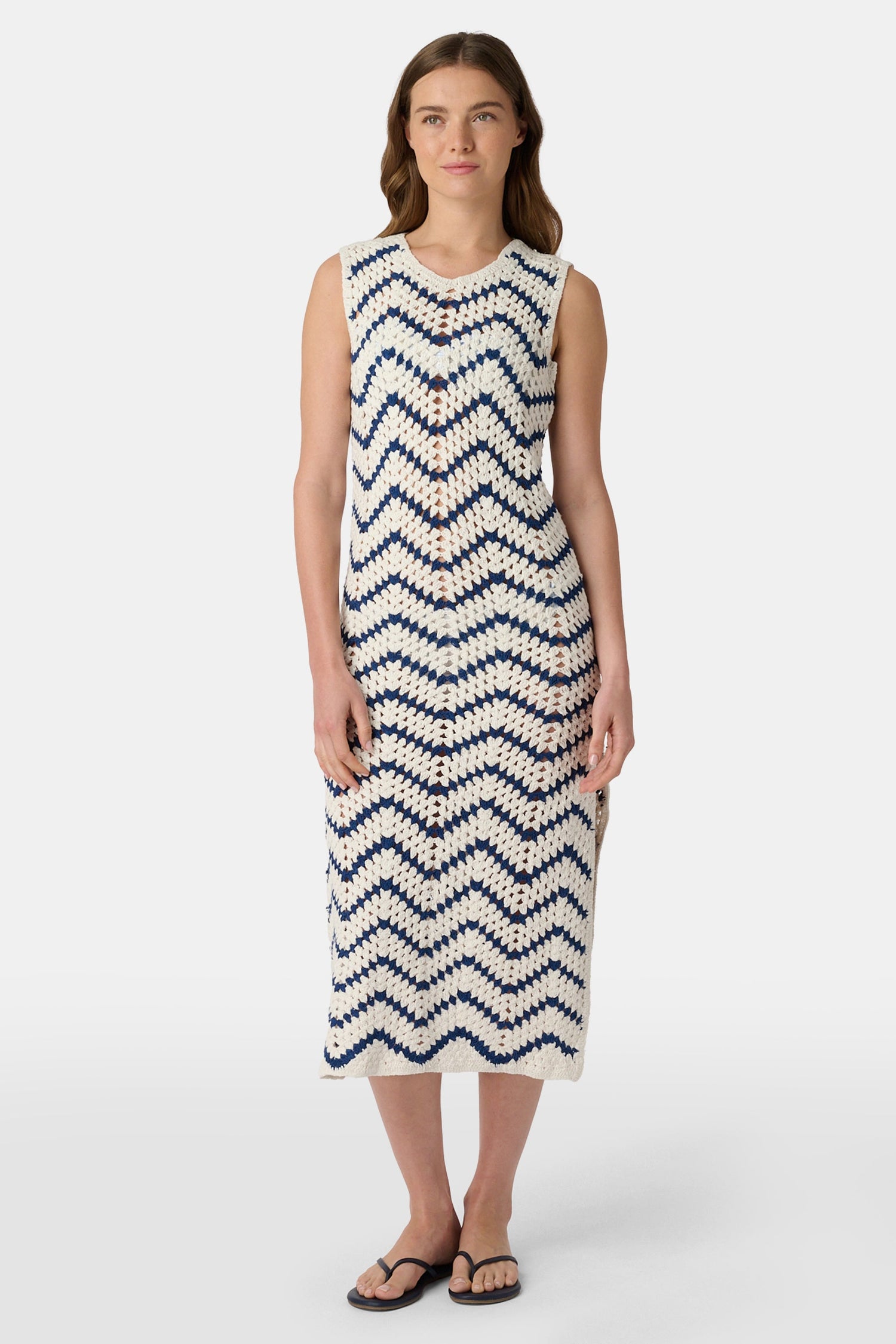 A woman wears the Roller Rabbit Zig Zag Irene Cover Up, a sleeveless, ankle-length crochet maxi dress with a blue and white zig zag pattern. She pairs it with black sandals and chunky striped bracelets, her straight brown hair worn loose.