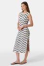 A woman wears the Roller Rabbit Zig Zag Irene Cover Up, a sleeveless, ankle-length crochet maxi dress with a blue and white zig zag pattern. She pairs it with black sandals and chunky striped bracelets, her straight brown hair worn loose.