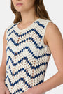 A woman wears the Roller Rabbit Zig Zag Irene Cover Up, a sleeveless, ankle-length crochet maxi dress with a blue and white zig zag pattern. She pairs it with black sandals and chunky striped bracelets, her straight brown hair worn loose.