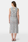A woman wears the Roller Rabbit Zig Zag Irene Cover Up, a sleeveless, ankle-length crochet maxi dress with a blue and white zig zag pattern. She pairs it with black sandals and chunky striped bracelets, her straight brown hair worn loose.
