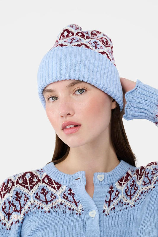 A young woman with fair skin and light makeup gazes at the camera, one hand on her hat, wearing a light blue Roller Rabbit Jemina Fairisle Beanie with a red and white pattern that matches her sweater.