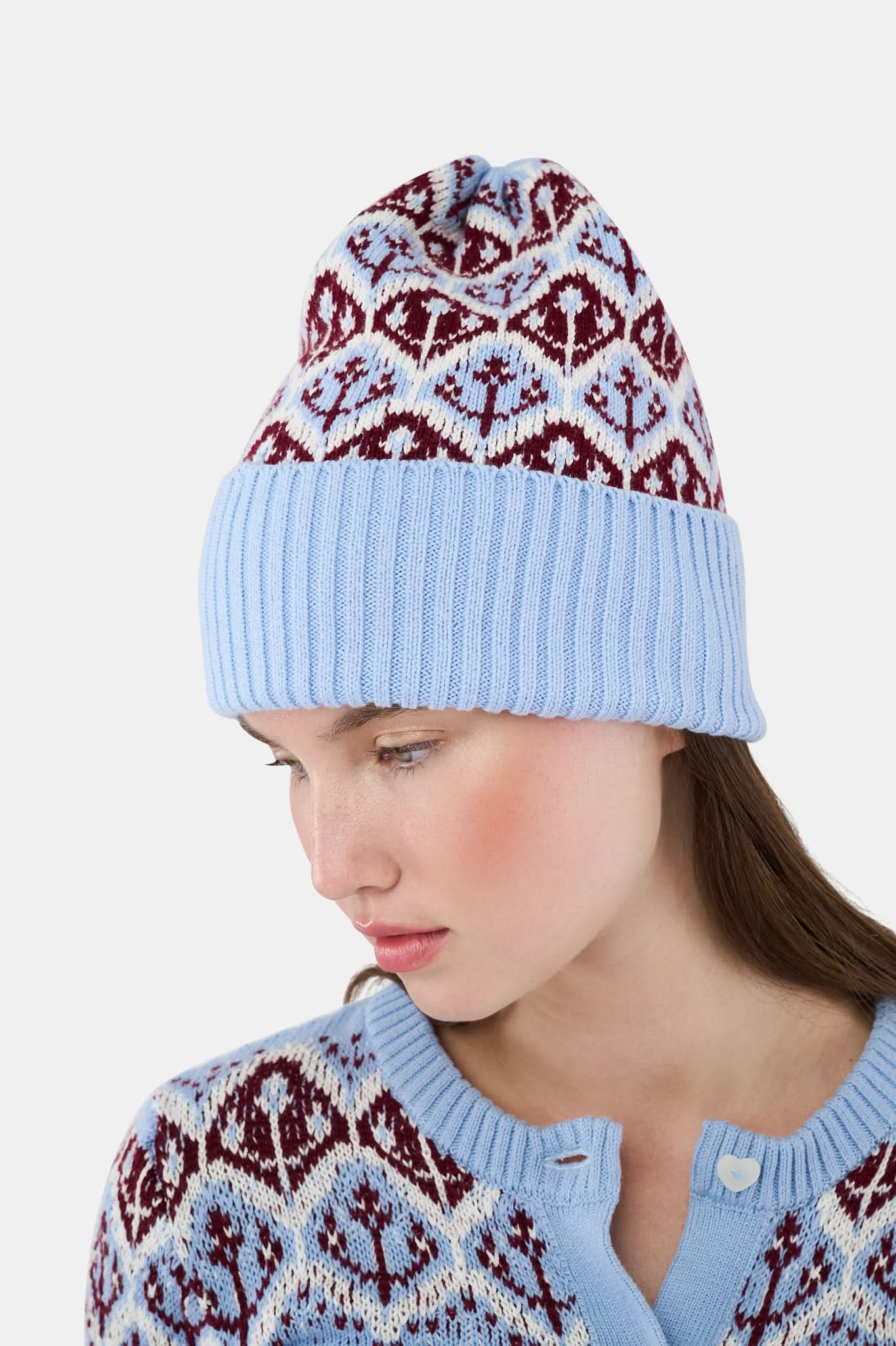 A young woman with fair skin and light makeup gazes at the camera, one hand on her hat, wearing a light blue Roller Rabbit Jemina Fairisle Beanie with a red and white pattern that matches her sweater.