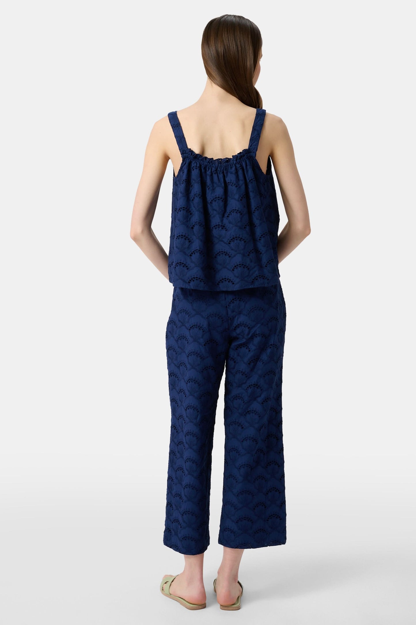 A woman with long brown hair wears the Roller Rabbit Jemina Eyelet Estelle Top in navy and wide-leg pants with a scalloped texture, paired with light green slide sandals. She stands against a plain white background.