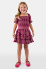 A young child wears the Roller Rabbit Kids Marquis Floral Bibi Skirt paired with maroon heart-accented shoes, standing against a plain white background.