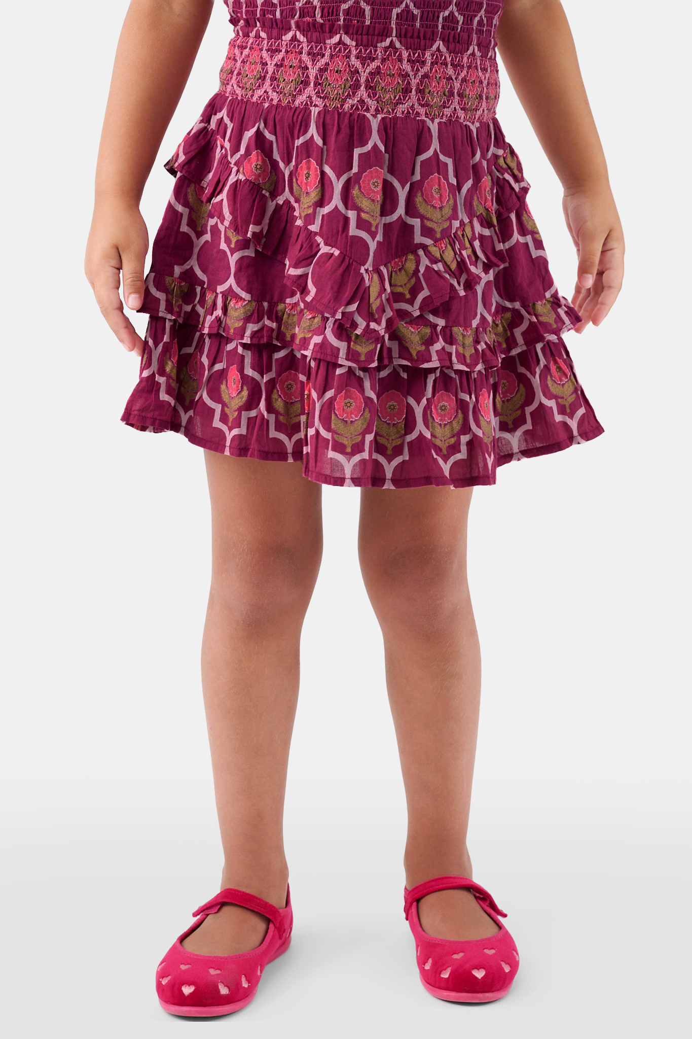 A young child wears the Roller Rabbit Kids Marquis Floral Bibi Skirt paired with maroon heart-accented shoes, standing against a plain white background.