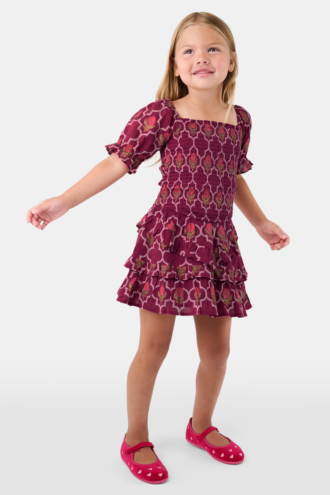 A young child wears the Roller Rabbit Kids Marquis Floral Bibi Skirt paired with maroon heart-accented shoes, standing against a plain white background.