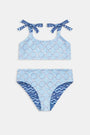 A girl in the Roller Rabbit Kids Fish Stripe/Ruban Reversible Cora Bikini stands by a pool, holding a striped towel behind her with outstretched arms. Her long hair is loose, and her swimwear offers UPF 50+ sun protection. Face partially out of frame.
