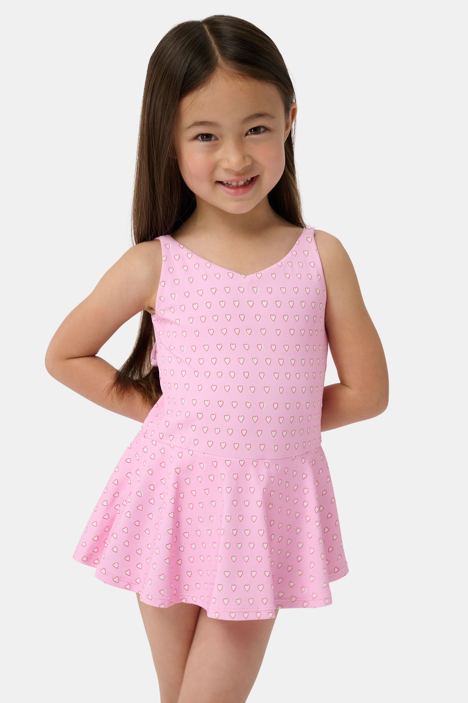 A young girl with long brown hair smiles confidently, wearing the Roller Rabbit Kids Hearts Dory One Piece swimsuit. She stands against a plain light gray background.