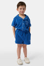 A child wears Roller Rabbit’s Kids Monkey Embroidery Hanni Short in navy with an embroidered figure on the left leg, matching blue top, and white velcro sneakers. Perfect summer shorts for active days.