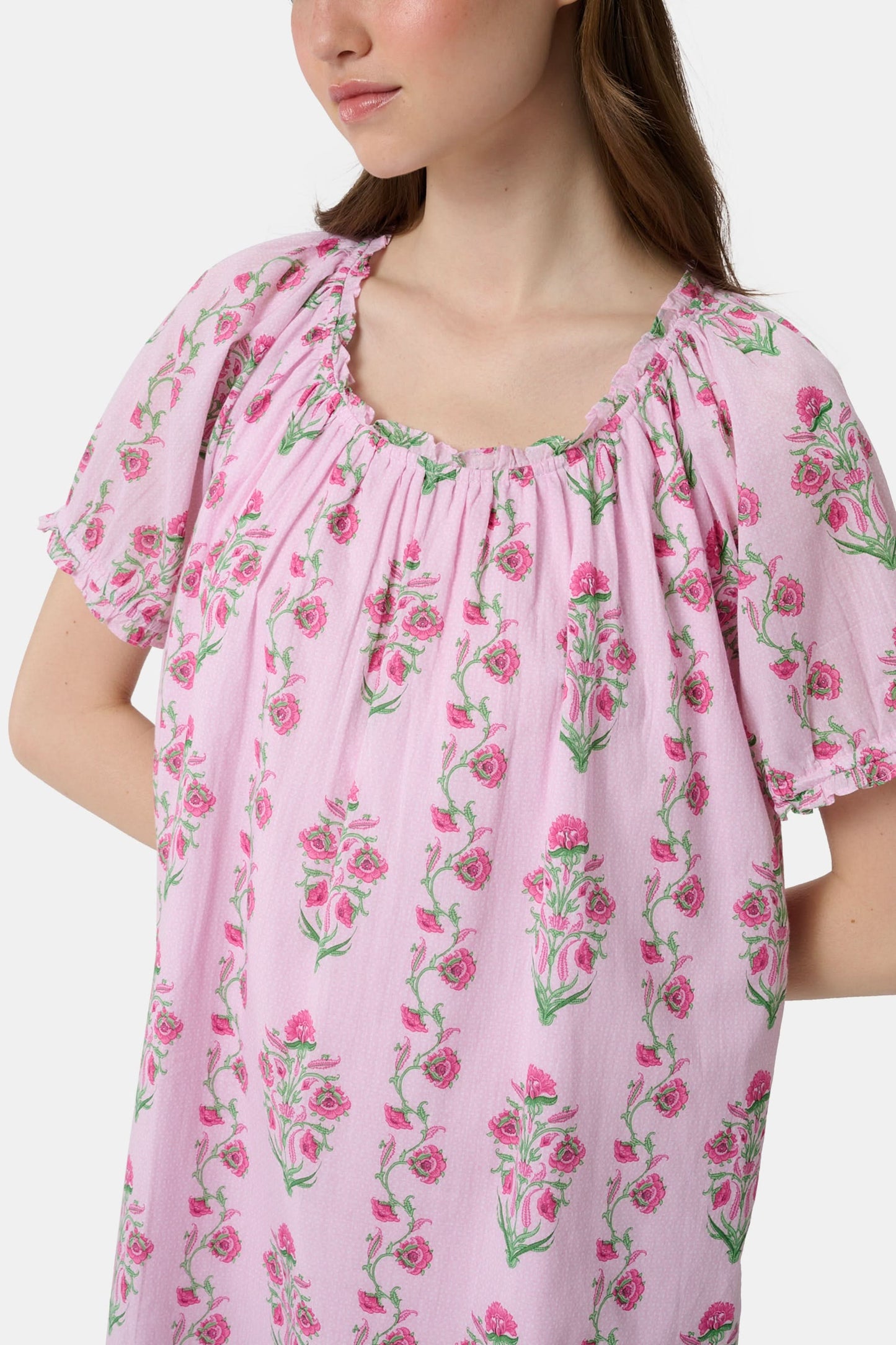 A woman with long brown hair wears the Roller Rabbit Lily Stripe Briella Dress, a loose cotton voile lounge dress with puffed short sleeves and a green and pink floral pattern, standing against a plain light background.