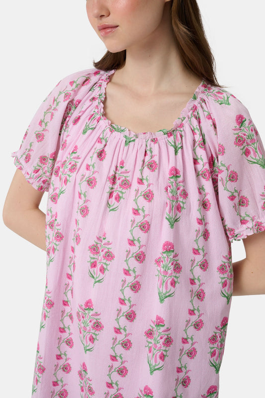 A woman with long brown hair wears the Roller Rabbit Lily Stripe Briella Dress, a loose cotton voile lounge dress with puffed short sleeves and a green and pink floral pattern, standing against a plain light background.
