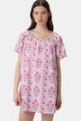 A woman with long brown hair wears the Roller Rabbit Lily Stripe Briella Dress, a loose cotton voile lounge dress with puffed short sleeves and a green and pink floral pattern, standing against a plain light background.