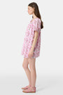 A woman with long brown hair wears the Roller Rabbit Lily Stripe Briella Dress, a loose cotton voile lounge dress with puffed short sleeves and a green and pink floral pattern, standing against a plain light background.