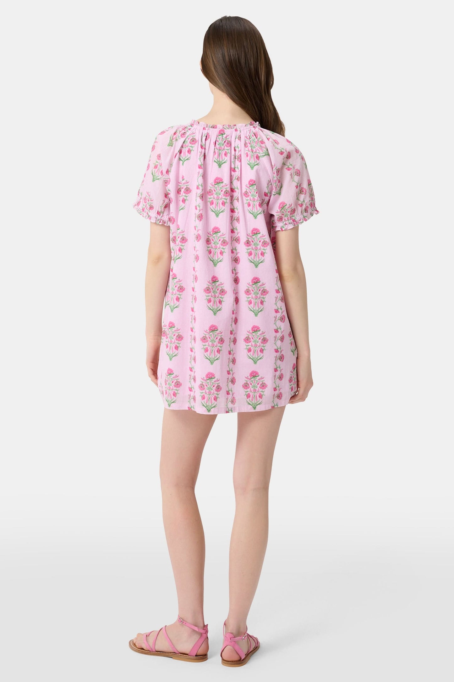 A woman with long brown hair wears the Roller Rabbit Lily Stripe Briella Dress, a loose cotton voile lounge dress with puffed short sleeves and a green and pink floral pattern, standing against a plain light background.