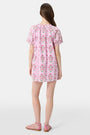 A woman with long brown hair wears the Roller Rabbit Lily Stripe Briella Dress, a loose cotton voile lounge dress with puffed short sleeves and a green and pink floral pattern, standing against a plain light background.