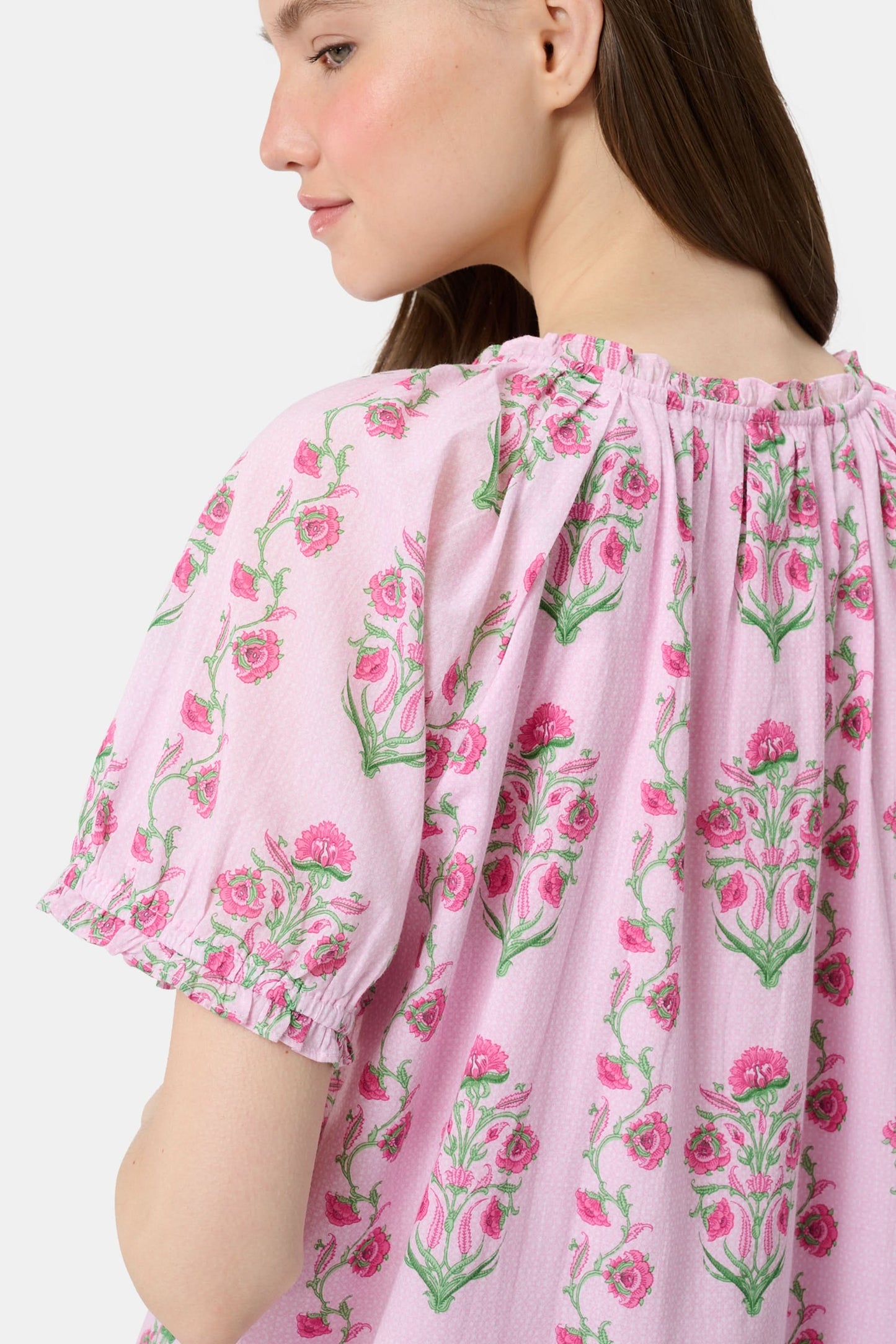 A woman with long brown hair wears the Roller Rabbit Lily Stripe Briella Dress, a loose cotton voile lounge dress with puffed short sleeves and a green and pink floral pattern, standing against a plain light background.