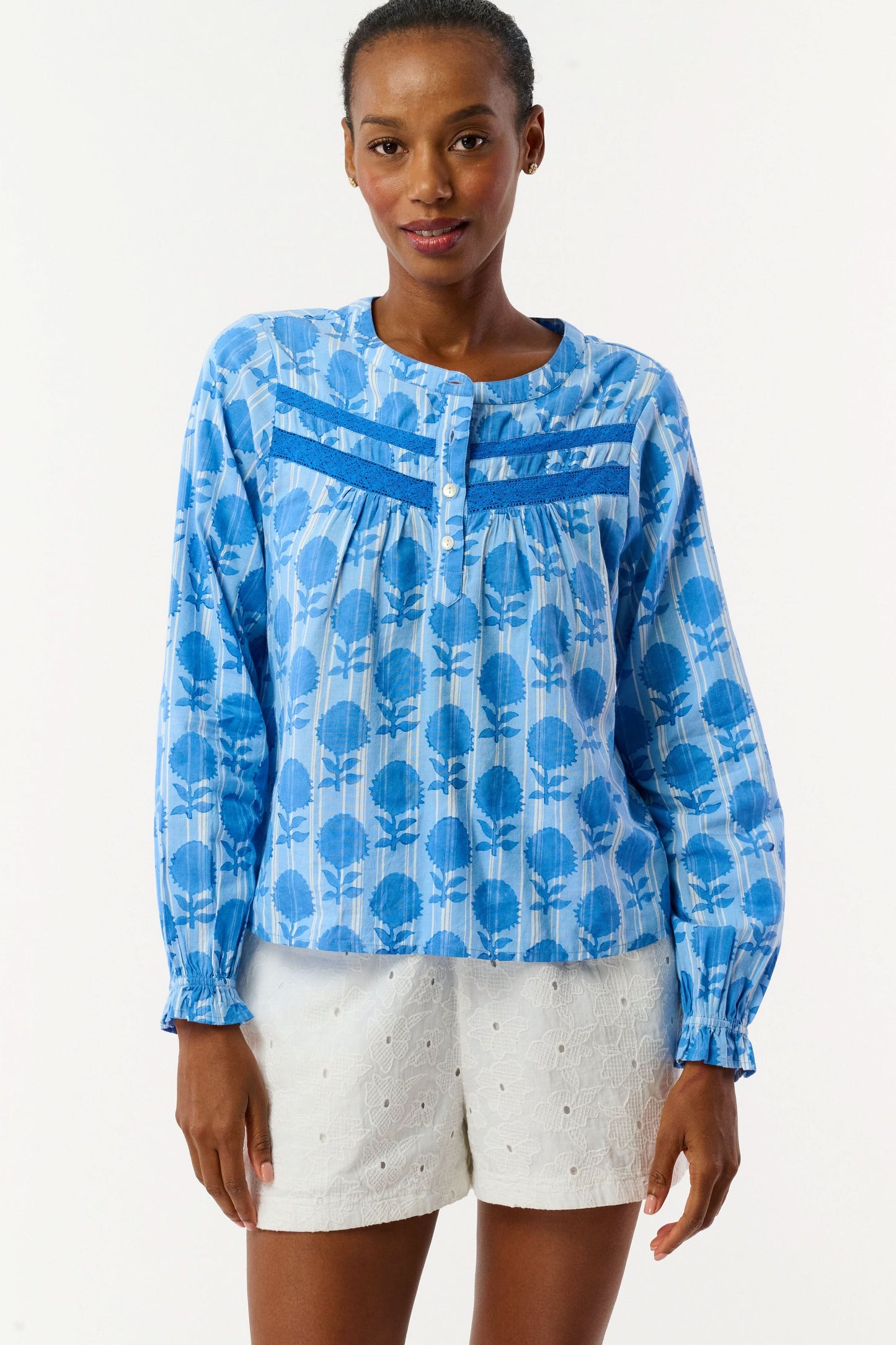A woman models the Linna Floral Jasper Top by Roller Rabbit, a blue long-sleeve blouse, paired with white shorts and poses against a plain white background, smiling gently at the camera.
