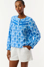 A woman models the Linna Floral Jasper Top by Roller Rabbit, a blue long-sleeve blouse, paired with white shorts and poses against a plain white background, smiling gently at the camera.