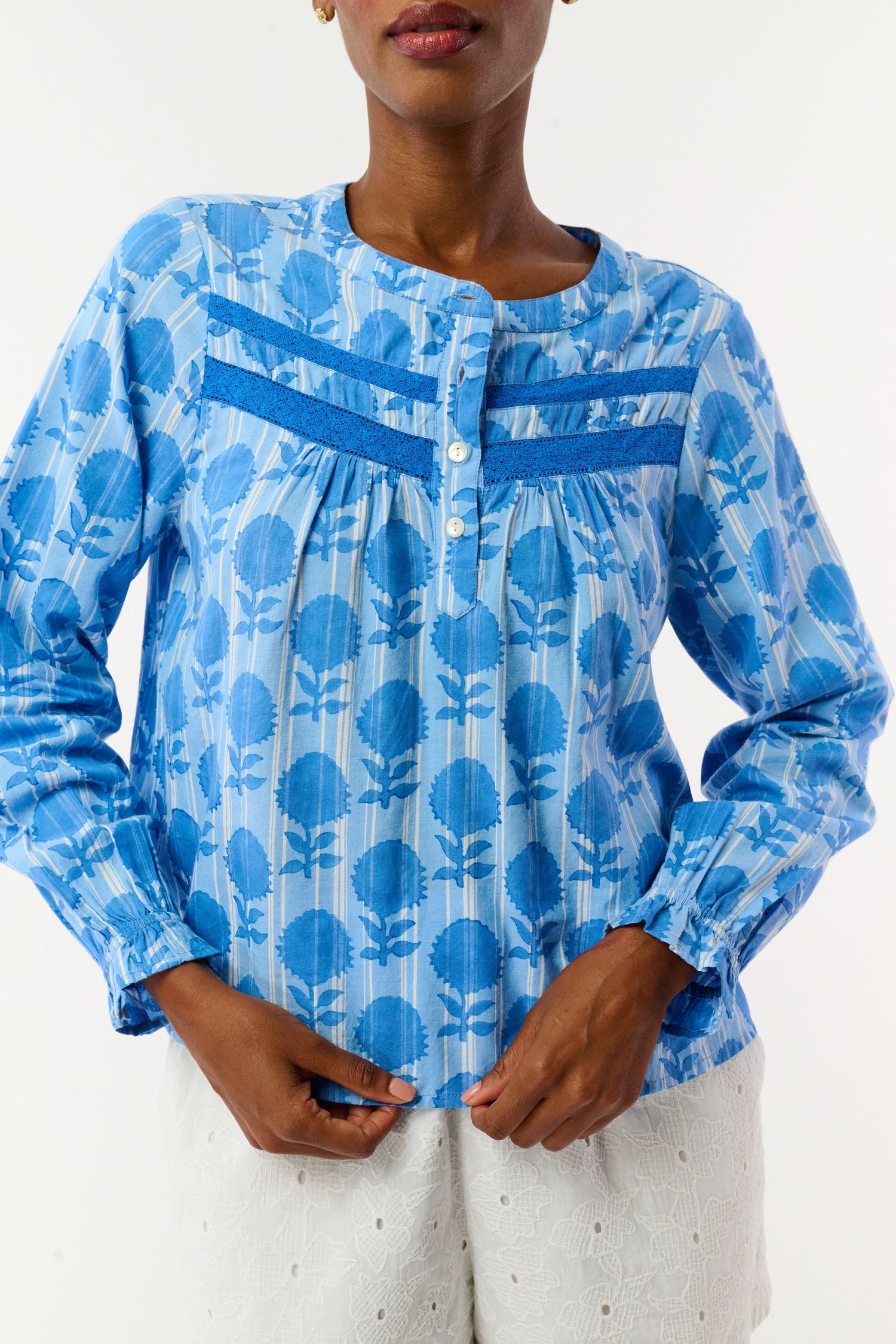 A woman models the Linna Floral Jasper Top by Roller Rabbit, a blue long-sleeve blouse, paired with white shorts and poses against a plain white background, smiling gently at the camera.