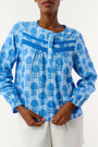 A woman models the Linna Floral Jasper Top by Roller Rabbit, a blue long-sleeve blouse, paired with white shorts and poses against a plain white background, smiling gently at the camera.