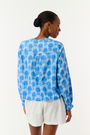 A woman models the Linna Floral Jasper Top by Roller Rabbit, a blue long-sleeve blouse, paired with white shorts and poses against a plain white background, smiling gently at the camera.