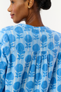 A woman models the Linna Floral Jasper Top by Roller Rabbit, a blue long-sleeve blouse, paired with white shorts and poses against a plain white background, smiling gently at the camera.