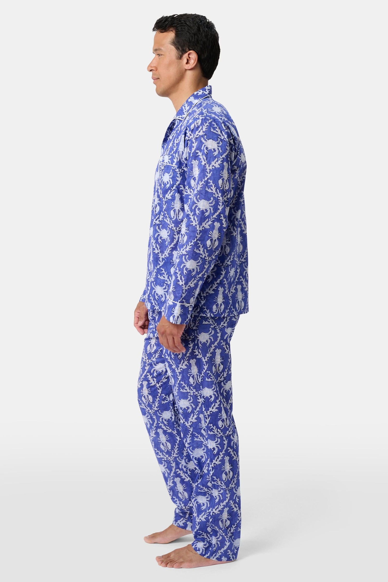 A man stands barefoot in Roller Rabbit’s Mens Lobster Trellis Loungewear, a matching blue and white floral cotton voile pajama set with a button-up long-sleeve shirt and pants. He has one hand in his pocket and looks forward.